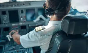 Pilot w słuchawkach i okularach siedzi w kokpicie samolotu, trzymając ręce na sterach.