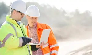 Dwóch mężczyzn w kaskach i kamizelkach odblaskowych analizuje dane na laptopie. To może być część ich kursu na kierownika budowy.