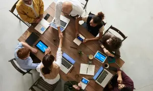 Grupa ludzi przy drewnianym stole, dwóch mężczyzn się ściska, laptopy i tablety.