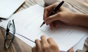 Dłoń pisząca długopisem na dokumencie, obok okulary i tablet.