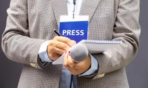 Dziennikarz w marynarce z identyfikatorem "PRESS" pisze w notatniku, trzymając mikrofon.