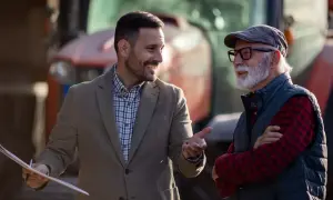 Dwóch mężczyzn rozmawia na tle traktora, jeden z nich trzyma dokumenty.