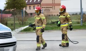 Dwóch strażaków w pełnym umundurowaniu, jeden z wężem wodnym, drugi z wężem.