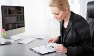 Kobieta w garniturze analizuje dokumenty za pomocą lupy przy biurku z komputerem.