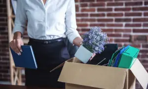 Kobieta w białej koszuli wkłada do kartonu doniczkę z kwiatami, książkę i segregatory.