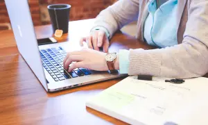 Kobieta pisze na laptopie, szukając odpowiedzi na pytanie jak znaleźć pracę. Na biurku leży też notatnik i kubek z herbatą.