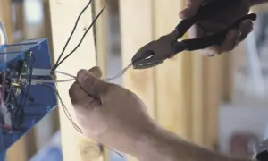 Dłonie trzymające kombinerki i przewody elektryczne przy niebieskiej puszce instalacyjnej.