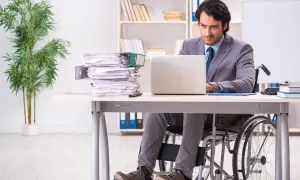 Mężczyzna na wózku inwalidzkim pracuje przy laptopie w biurze, obok stos dokumentów.
