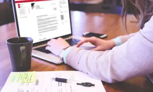 Kobieta pisze list motywacyjny na laptopie, obok kubek i notatki.