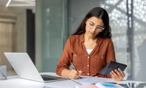 Kobieta w okularach liczy na kalkulatorze i pisze na dokumentach przy laptopie.