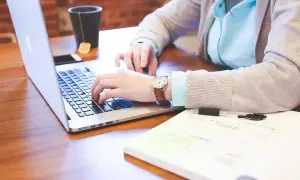 Osoba pisząca na laptopie, obok notatnik i kubek z herbatą.