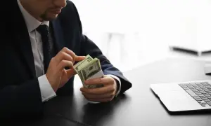 Mężczyzna w garniturze liczy banknoty dolarów przy biurku z laptopem.