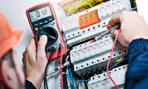 Elektryk w pomarańczowym kasku mierzy napięcie multimetrem w skrzynce z bezpiecznikami.