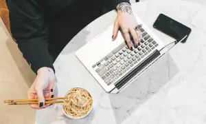 Osoba z tatuażami pracuje na laptopie, jedząc makaron pałeczkami.