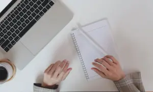 Dłonie nad notatnikiem i laptopem, filiżanka kawy.