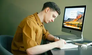 Mężczyzna w żółtej koszuli siedzi przy biurku i pisze w notatniku, przed nim komputer.