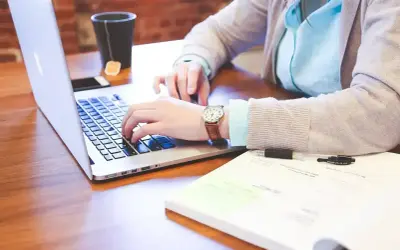 Osoba pisząca na laptopie, obok notatnik i kubek z herbatą.