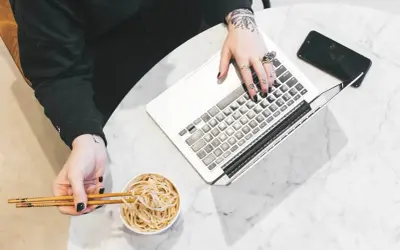 Osoba z tatuażami pracuje na laptopie, jedząc makaron pałeczkami.