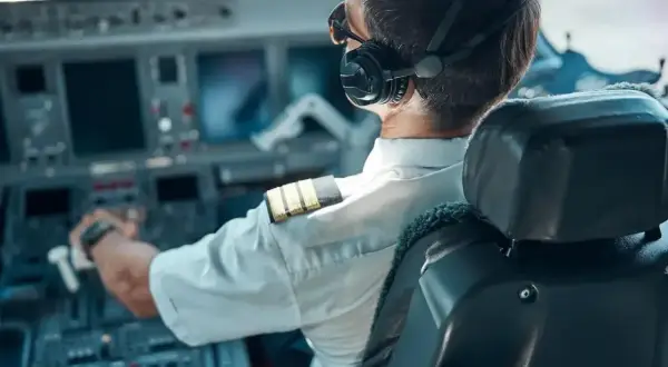 Pilot w słuchawkach i okularach siedzi w kokpicie samolotu, trzymając ręce na sterach.