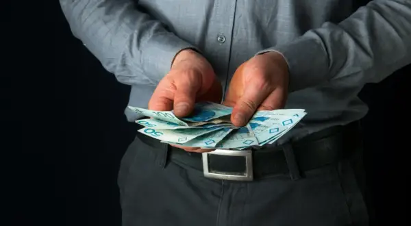 Mężczyzna w szarej koszuli trzyma w dłoniach polskie banknoty 50 i 100 zł.