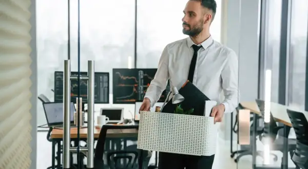 Mężczyzna w białej koszuli i krawacie niesie pudełko z rzeczami z biura.