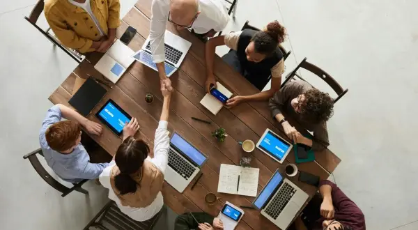 Grupa ludzi przy drewnianym stole, dwóch mężczyzn się ściska, laptopy i tablety.
