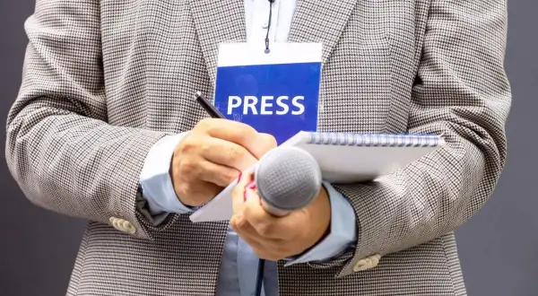 Dziennikarz w marynarce z identyfikatorem "PRESS" pisze w notatniku, trzymając mikrofon.