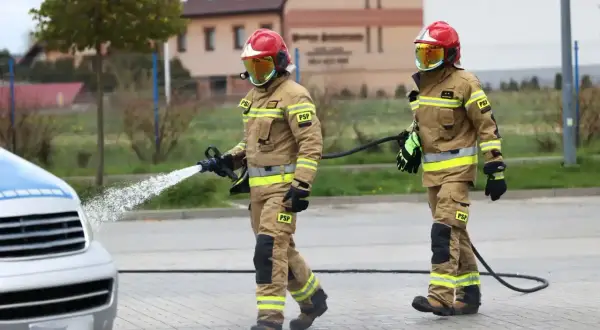 Dwóch strażaków w pełnym umundurowaniu, jeden z wężem wodnym, drugi z wężem.