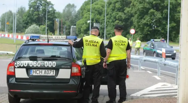 Dwóch funkcjonariuszy Służby Celnej w kamizelkach odblaskowych stoi przy oznakowanym radiowozie.