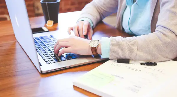 Kobieta pisze na laptopie, szukając odpowiedzi na pytanie jak znaleźć pracę. Na biurku leży też notatnik i kubek z herbatą.