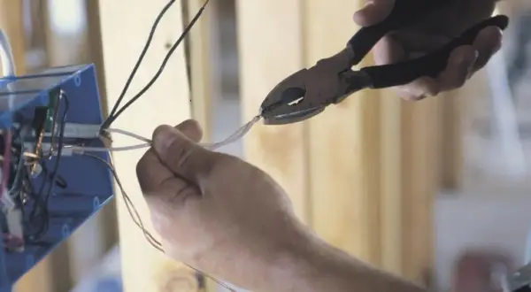 Dłonie trzymające kombinerki i przewody elektryczne przy niebieskiej puszce instalacyjnej.