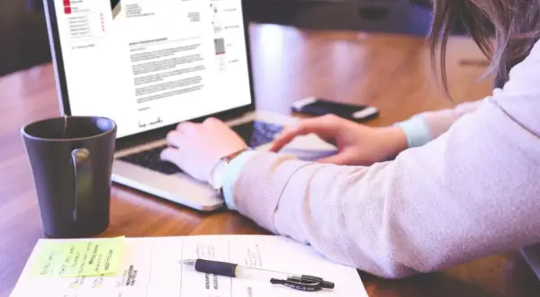 Kobieta pisze list motywacyjny na laptopie, obok kubek i notatki.