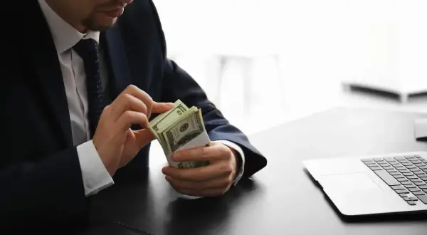 Mężczyzna w garniturze liczy banknoty dolarów przy biurku z laptopem.