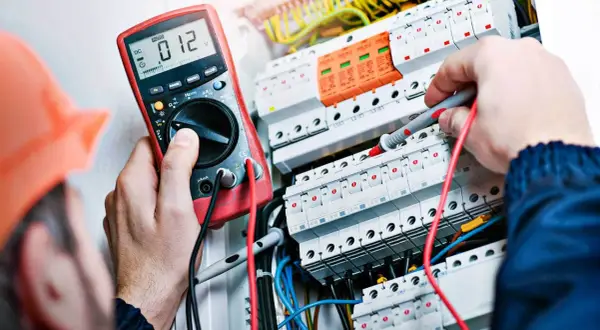 Elektryk w pomarańczowym kasku mierzy napięcie multimetrem w skrzynce z bezpiecznikami.