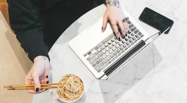 Osoba z tatuażami pracuje na laptopie, jedząc makaron pałeczkami.