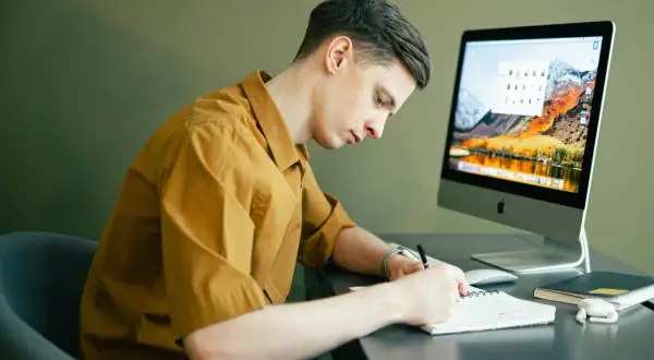 Mężczyzna w żółtej koszuli siedzi przy biurku i pisze w notatniku, przed nim komputer.