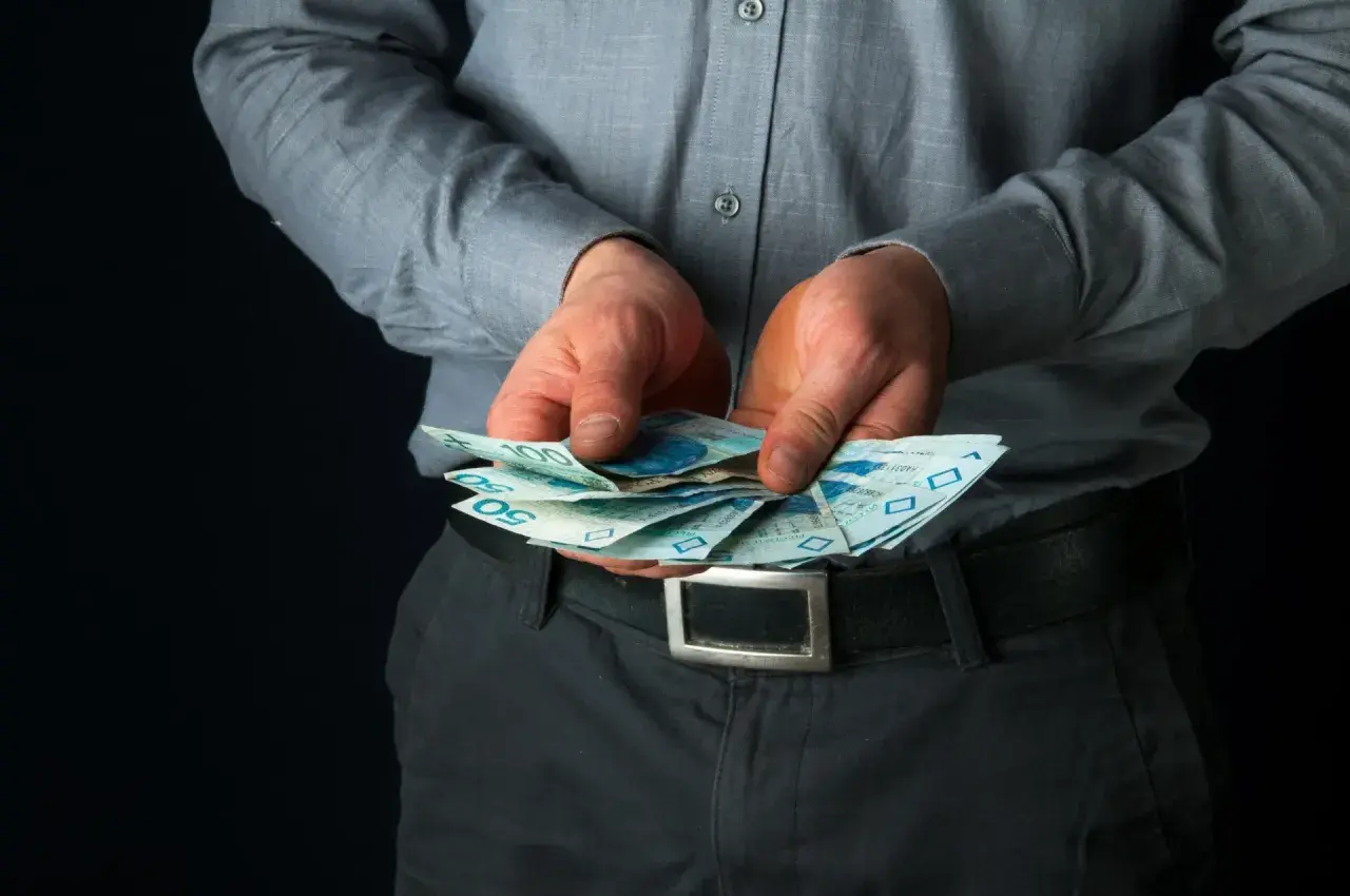 Mężczyzna w szarej koszuli trzyma w dłoniach polskie banknoty 50 i 100 zł.