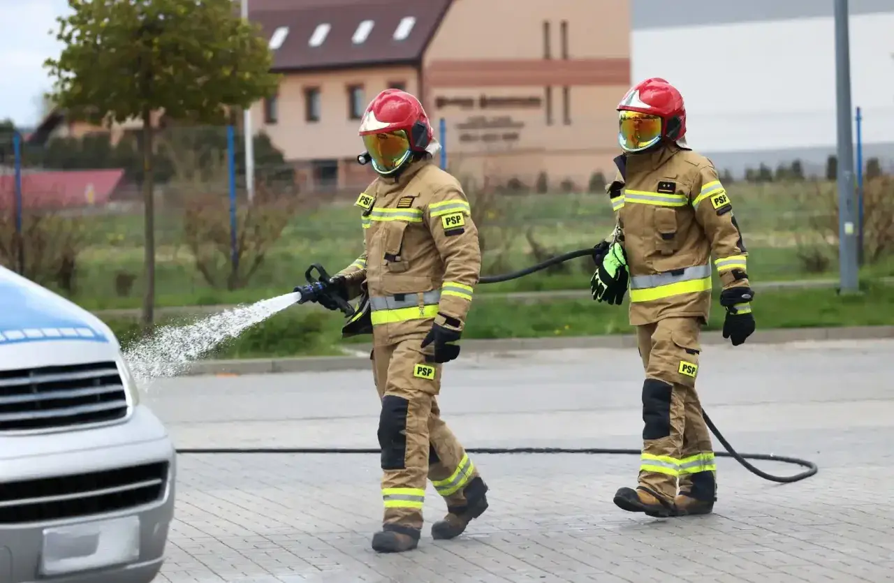 Dwóch strażaków w pełnym umundurowaniu, jeden z wężem wodnym, drugi z wężem.