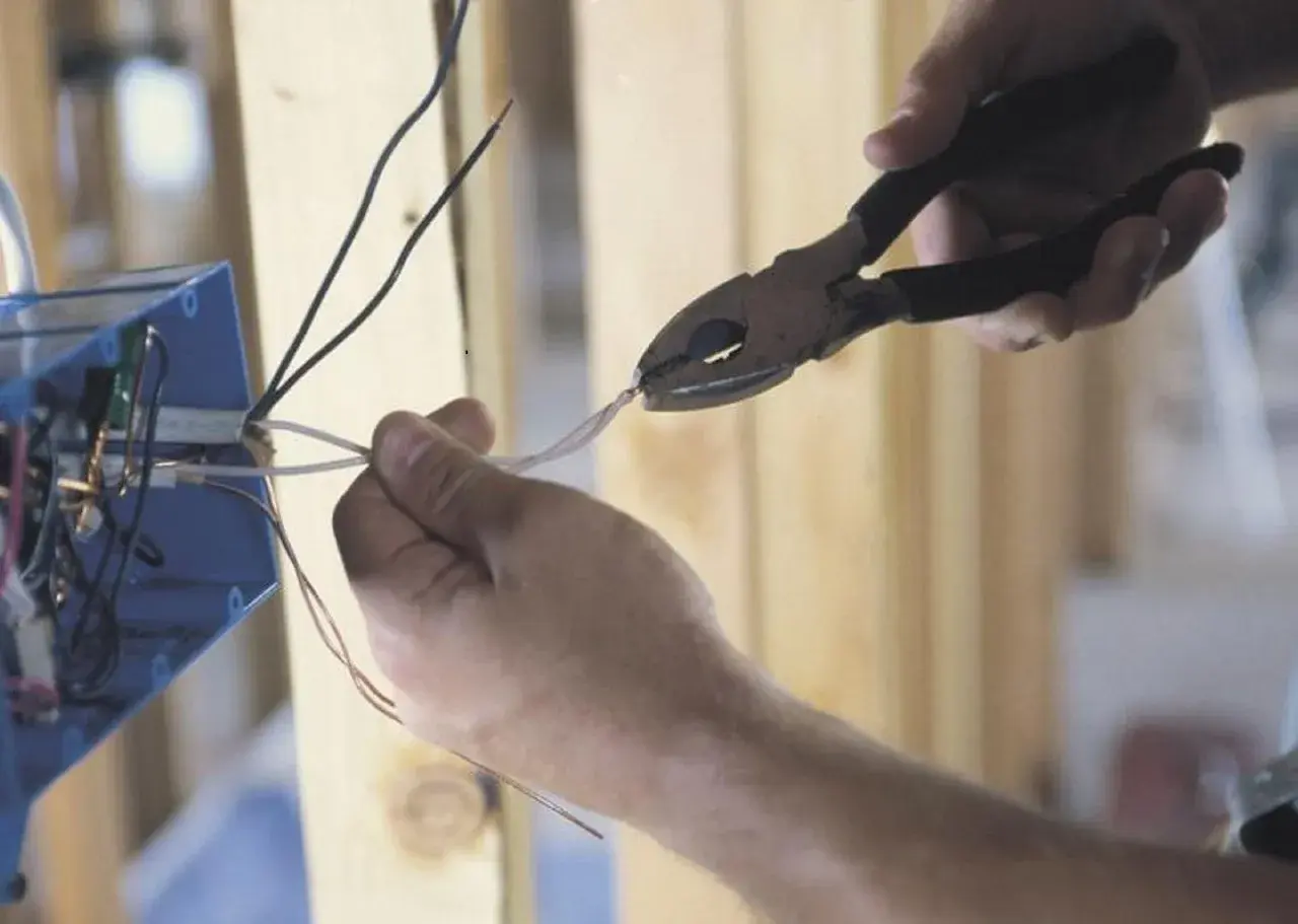 Dłonie trzymające kombinerki i przewody elektryczne przy niebieskiej puszce instalacyjnej.