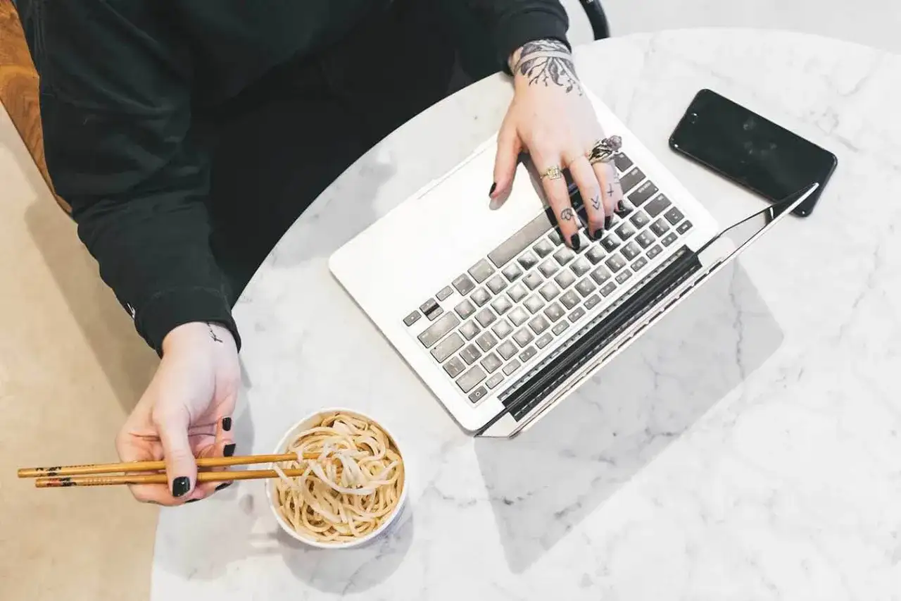 Osoba z tatuażami pracuje na laptopie, jedząc makaron pałeczkami.