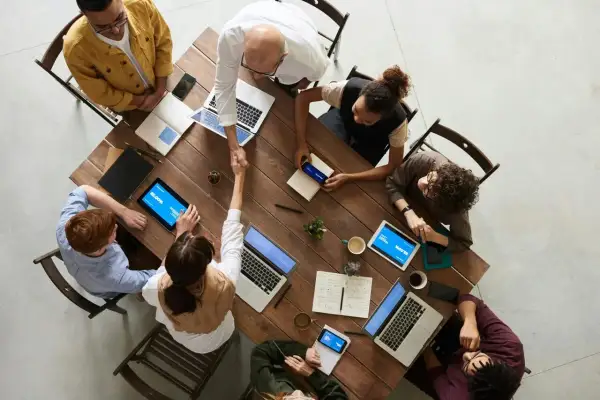 Grupa ludzi przy drewnianym stole, dwóch mężczyzn się ściska, laptopy i tablety.