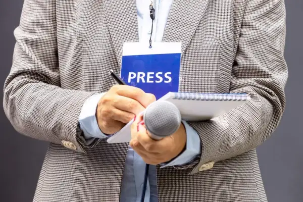 Dziennikarz w marynarce z identyfikatorem "PRESS" pisze w notatniku, trzymając mikrofon.