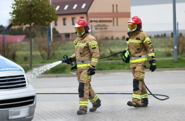 Dwóch strażaków w pełnym umundurowaniu, jeden z wężem wodnym, drugi z wężem.