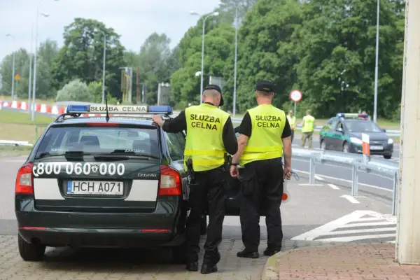 Dwóch funkcjonariuszy Służby Celnej w kamizelkach odblaskowych stoi przy oznakowanym radiowozie.