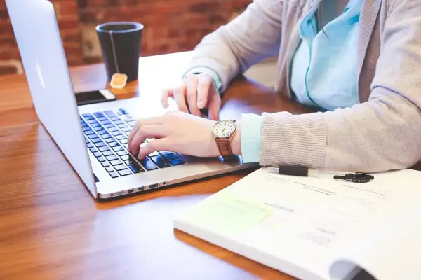 Kobieta pisze na laptopie, szukając odpowiedzi na pytanie jak znaleźć pracę. Na biurku leży też notatnik i kubek z herbatą.
