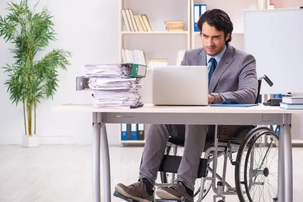 Mężczyzna na wózku inwalidzkim pracuje przy laptopie w biurze, obok stos dokumentów.