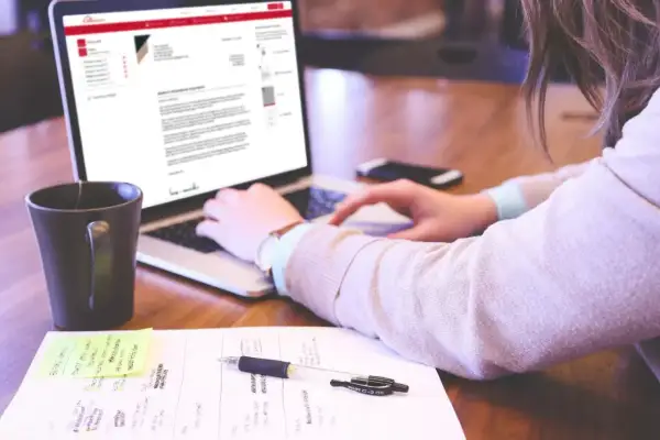 Kobieta pisze list motywacyjny na laptopie, obok kubek i notatki.