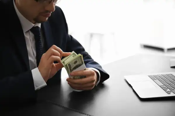 Mężczyzna w garniturze liczy banknoty dolarów przy biurku z laptopem.