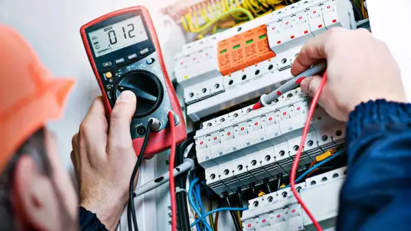 Elektryk w pomarańczowym kasku mierzy napięcie multimetrem w skrzynce z bezpiecznikami.